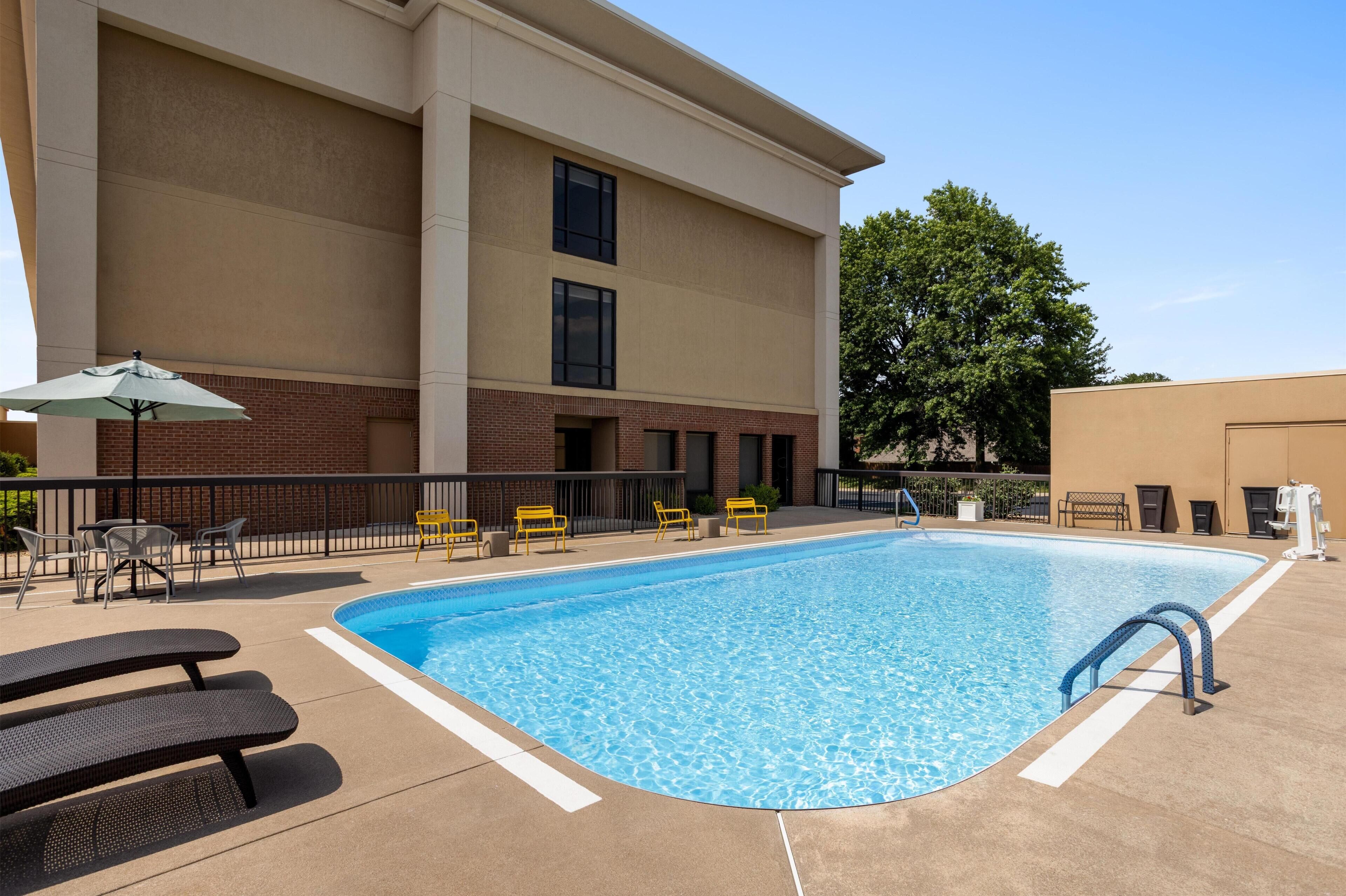 A rectangular outdoor swimming pool with a clear blue water surface, surrounded by a concrete deck. There are several yellow chairs and a white umbrella providing shade.