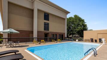 A rectangular outdoor swimming pool with a clear blue water surface, surrounded by a concrete deck. There are several yellow chairs and a white umbrella providing shade.