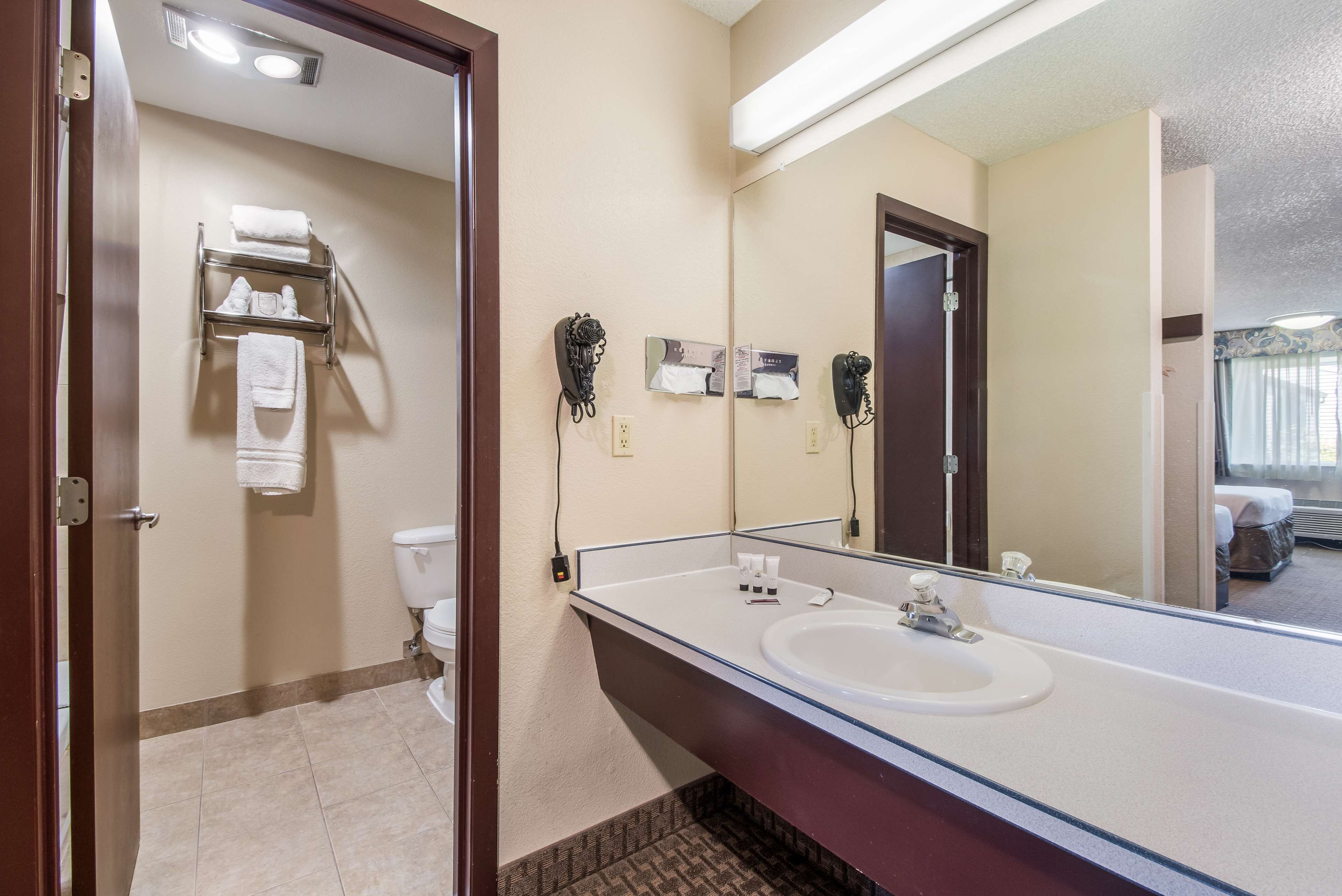 A hotel bathroom with a large mirror, a sink, and a toilet. There are towels on a rack and a hairdryer on the wall.