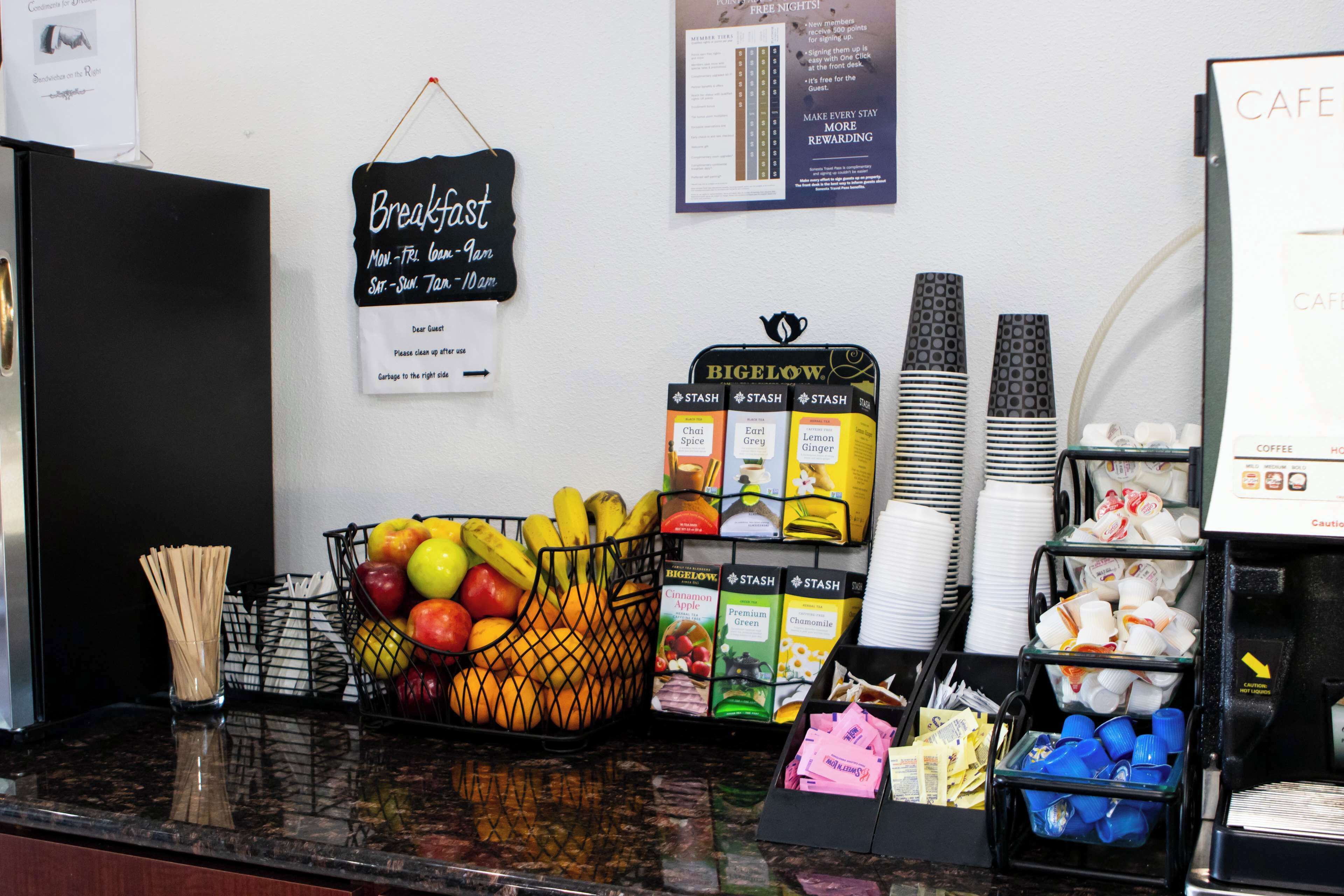 A breakfast counter with a sign indicating hours of operation, a basket of fruits, a coffee machine, and various coffee pods.