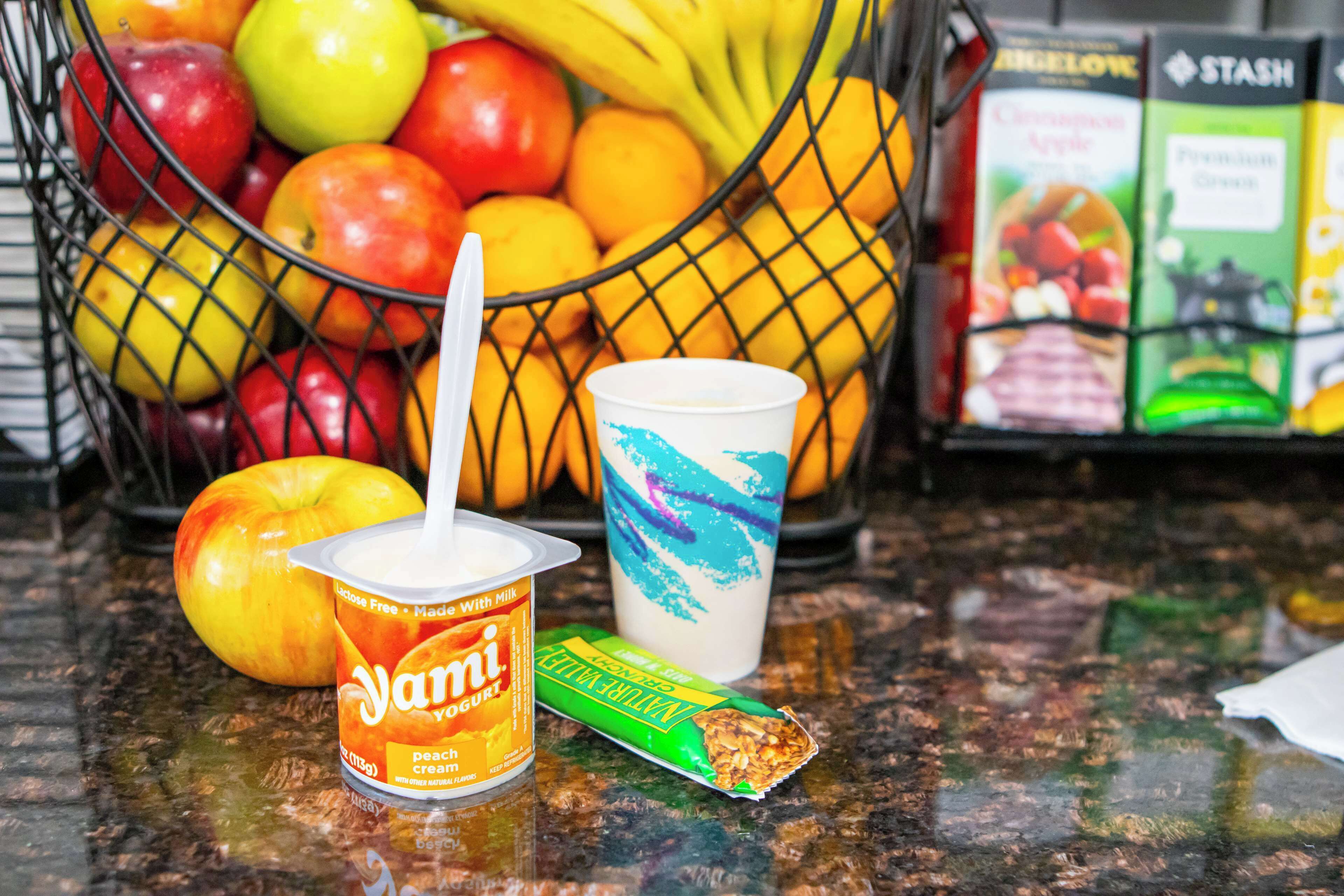 A fruit basket containing apples and oranges, a yogurt cup, and a granola bar on a granite countertop.