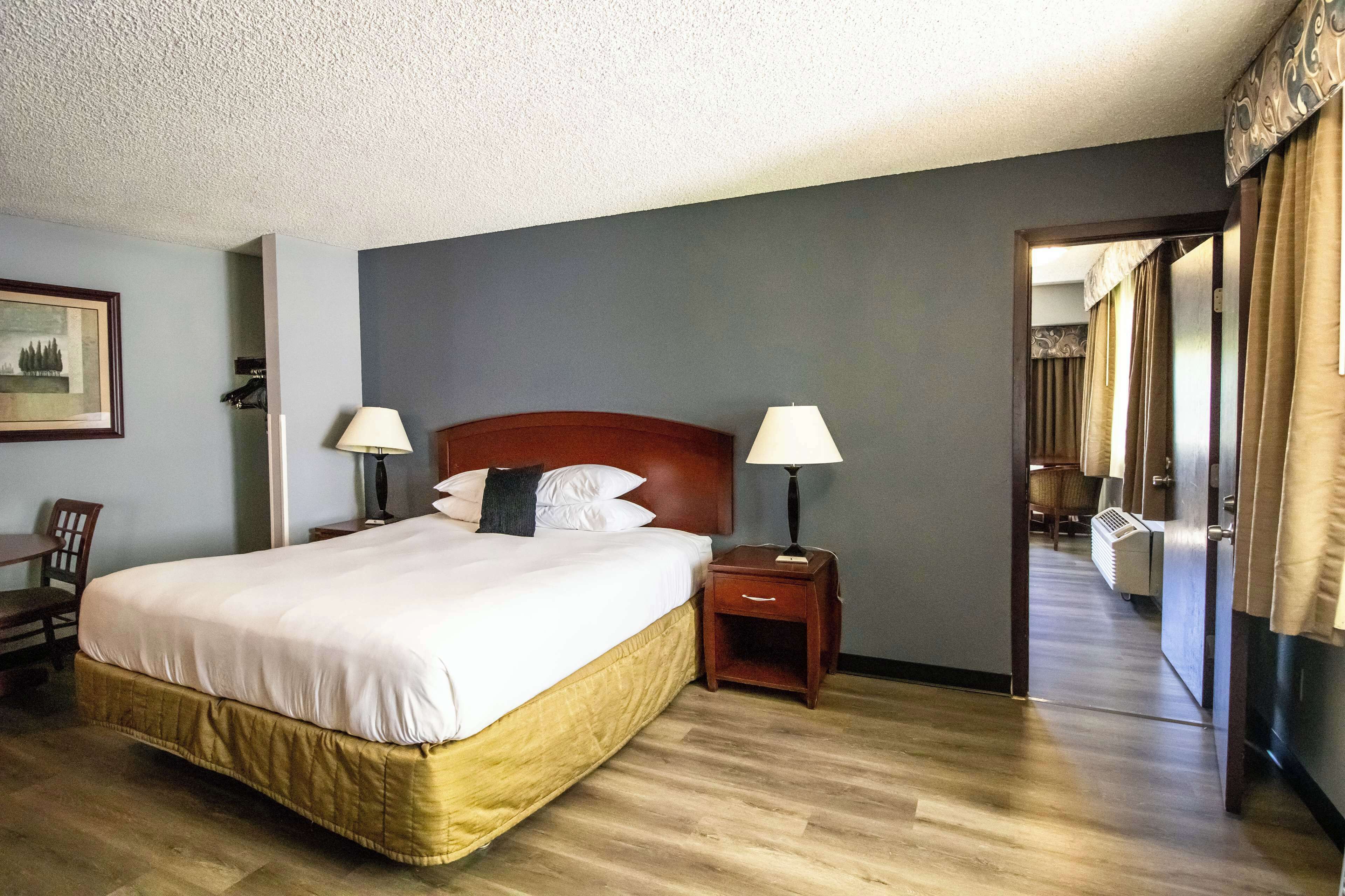 A hotel room with a large bed, two bedside tables with lamps, a wooden headboard, a framed picture on the wall, and a view through an open door.