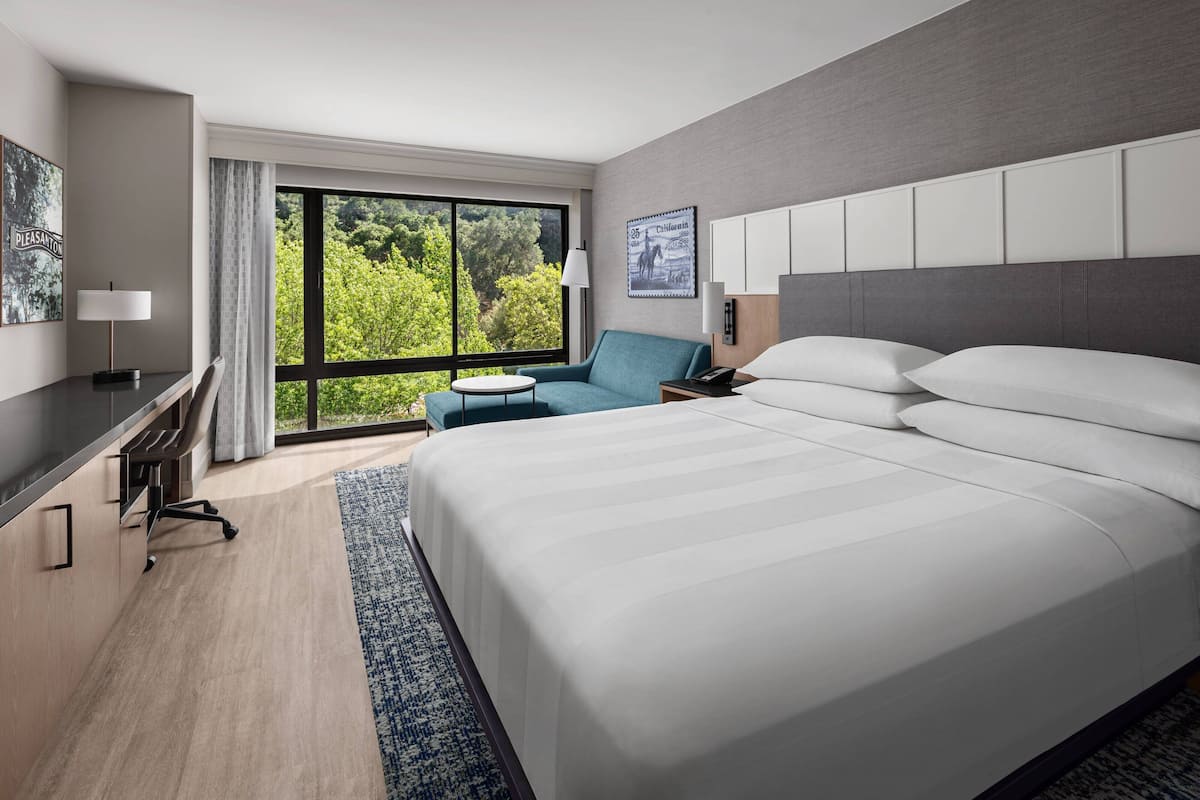 A modern hotel room with a large bed, a desk, a blue armchair, and a view of greenery through the window.