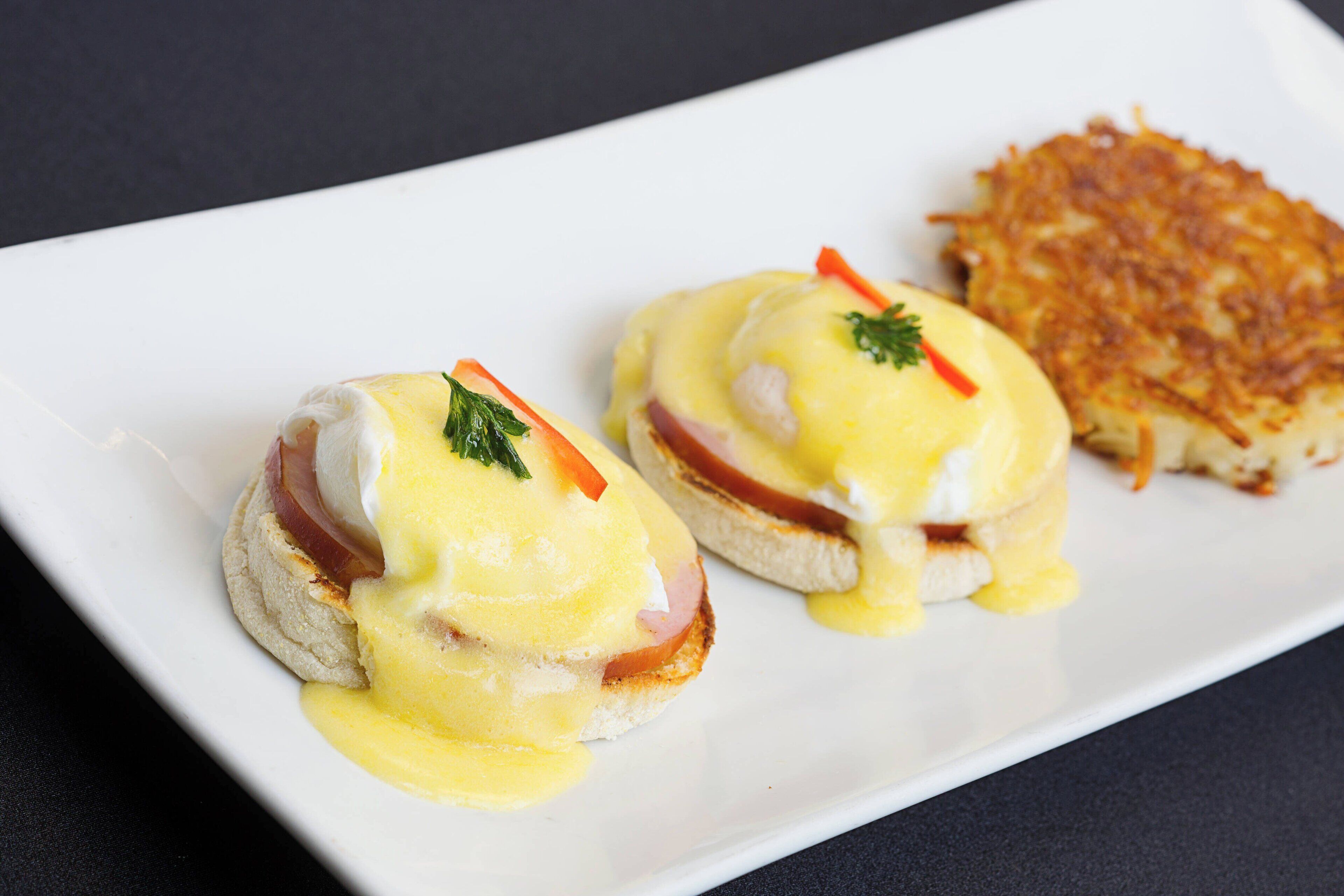 Two eggs benedict sandwiches on a white plate with hollandaise sauce and garnishes, and a side of hash browns.