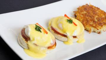 Two eggs benedict sandwiches on a white plate with hollandaise sauce and garnishes, and a side of hash browns.