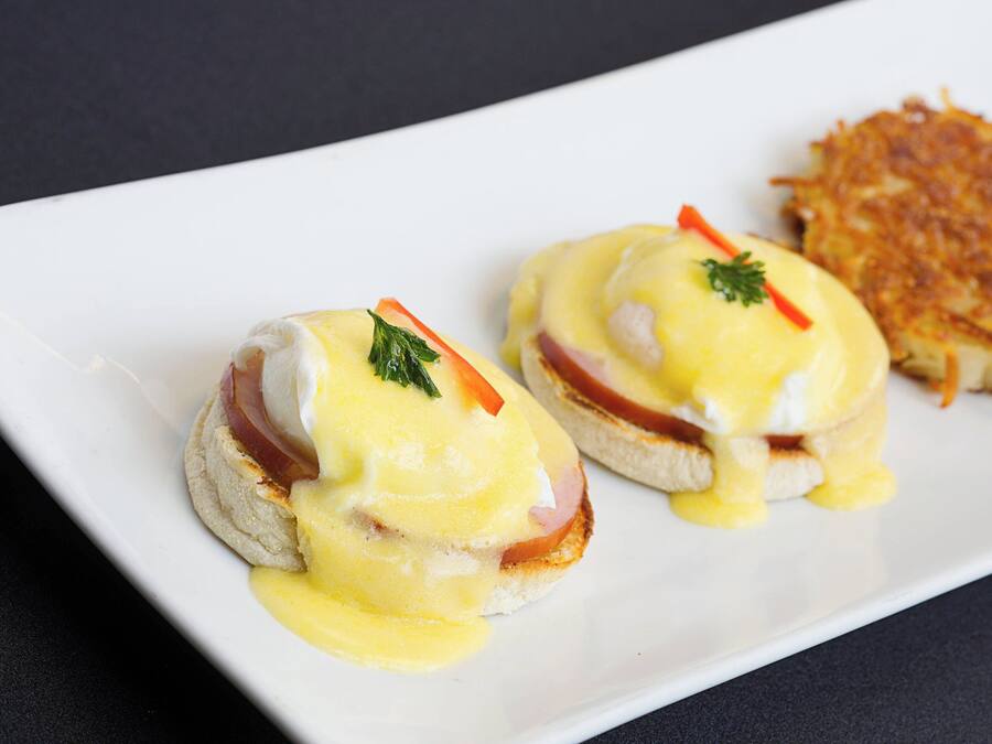 Two eggs benedict sandwiches on a white plate with hollandaise sauce and garnishes, and a side of hash browns.