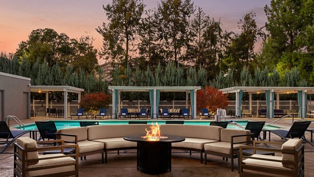 Zona junto a la piscina con una chimenea, sillones de descanso y un área de estar cubierta con vistas a árboles y al atardecer.