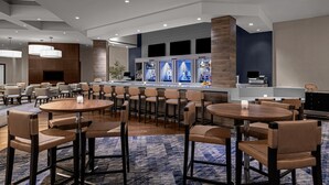 Un moderno vestíbulo de hotel con mesas y sillas redondas, una zona de bar con refrigeradores y una planta grande.