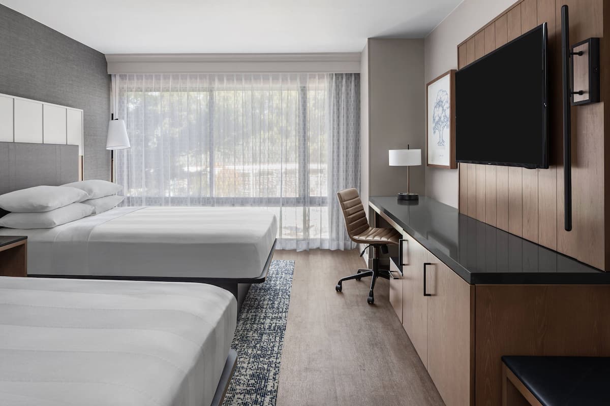 A hotel room with two beds, a desk, a chair, a TV, and a large window with sheer curtains.