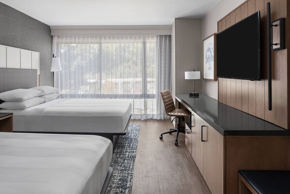 A hotel room with two beds, a desk, a chair, a TV, and a large window with sheer curtains.