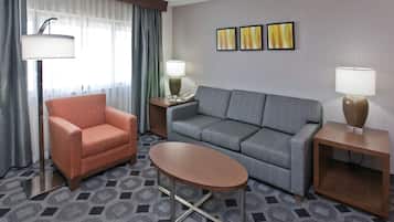 A hotel room with a grey sofa, an orange armchair, a small round table, and two bedside tables with lamps.