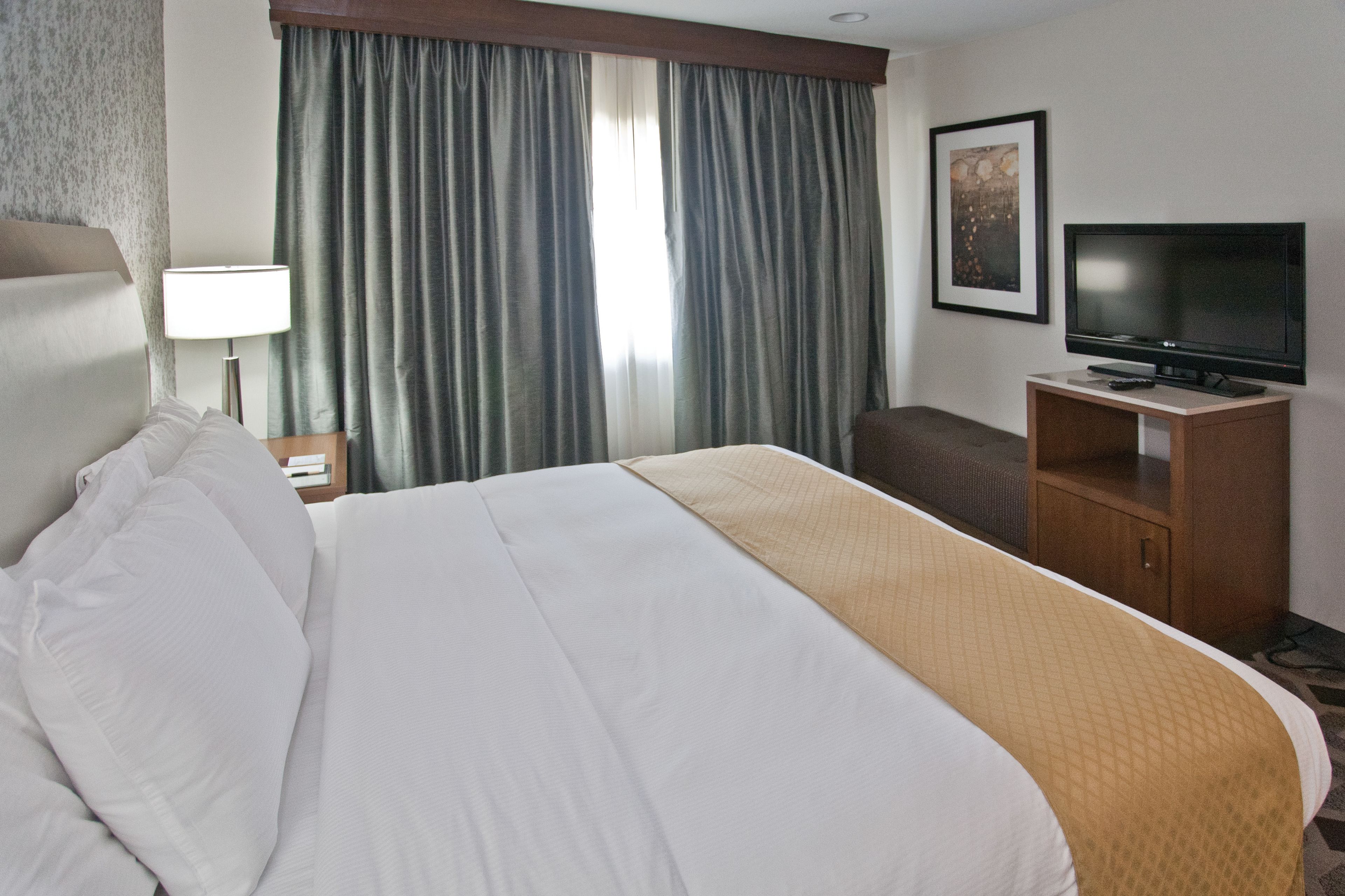 A hotel room with a large bed, a television on a wooden stand, a desk with a lamp, and a window with curtains.