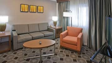 A hotel room with a grey sofa, an orange armchair, a round coffee table, and a TV on a stand.