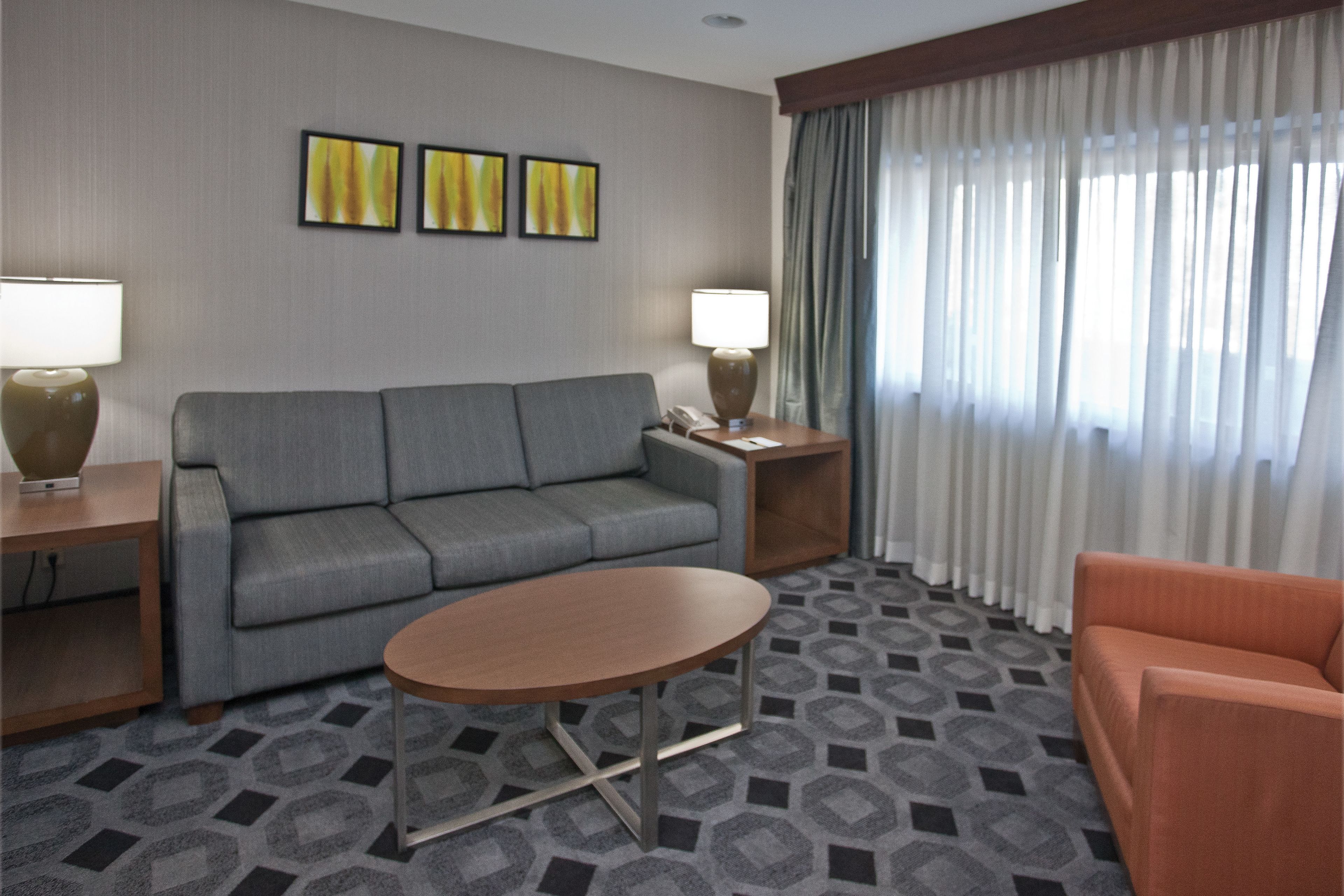 Ein Hotelzimmer mit grauem Sofa, einem runden Tisch, einem kleinen orangefarbenen Sessel, einer Lampe, einem Telefon und einem Fenster mit durchsichtigen Vorhängen.