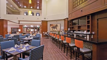 A spacious hotel lobby with multiple dining areas, a bar, and a high ceiling with wooden paneling.