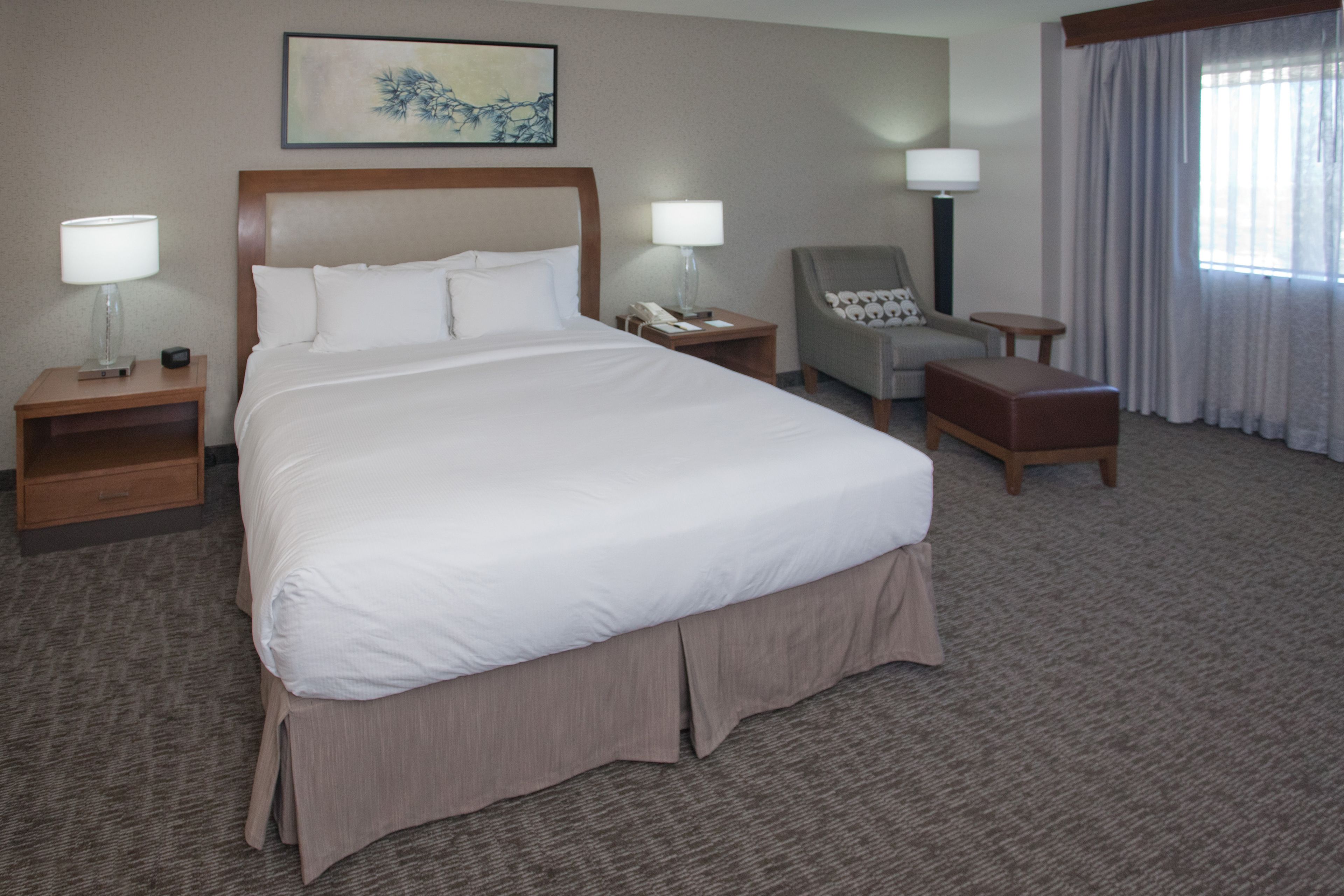 Une chambre d’hôtel avec un grand lit, deux tables de chevet avec des lampes, une chaise, une petite table et une fenêtre avec des rideaux.