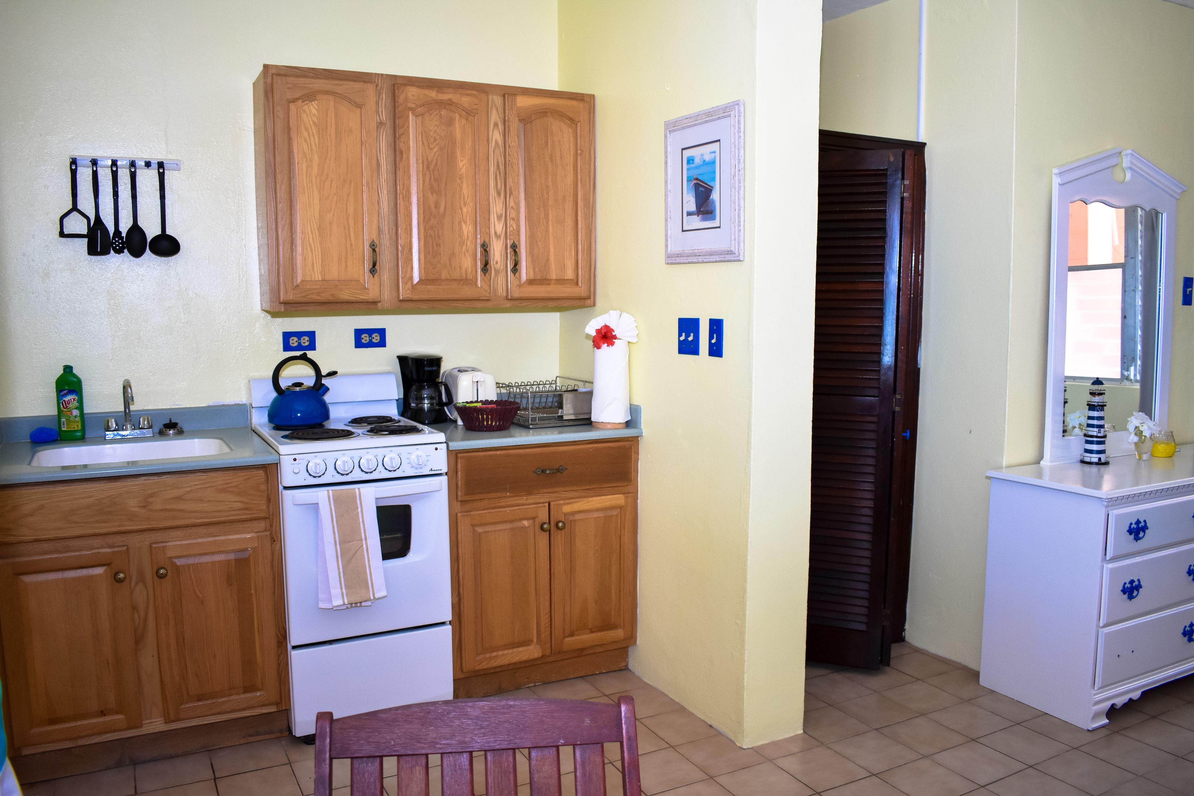 A compact kitchen with wooden cabinets, a stove, and a sink. There is a chair and a small table.