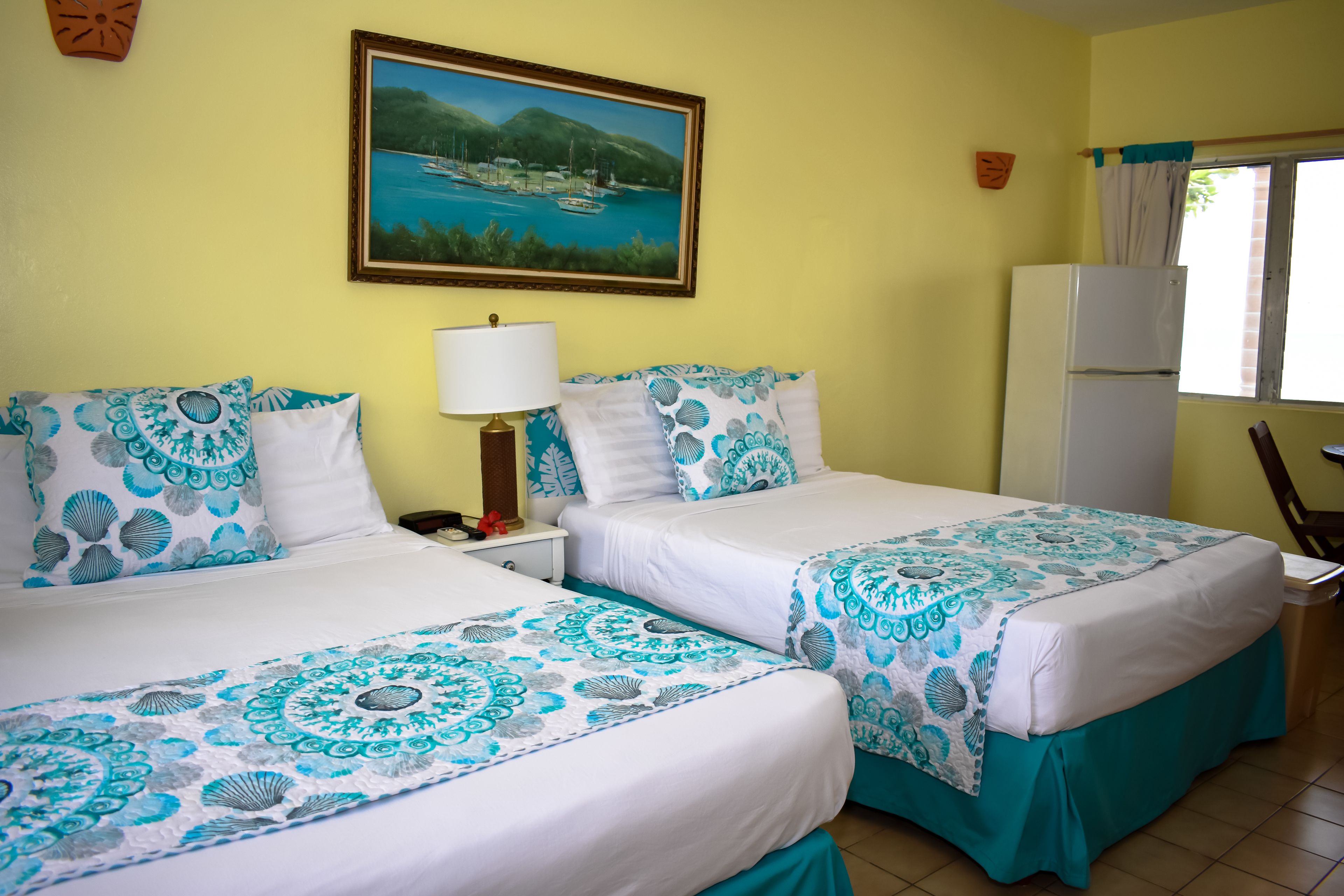 A hotel room with two beds, a painting of a coastal scene, a refrigerator, and a window with curtains.