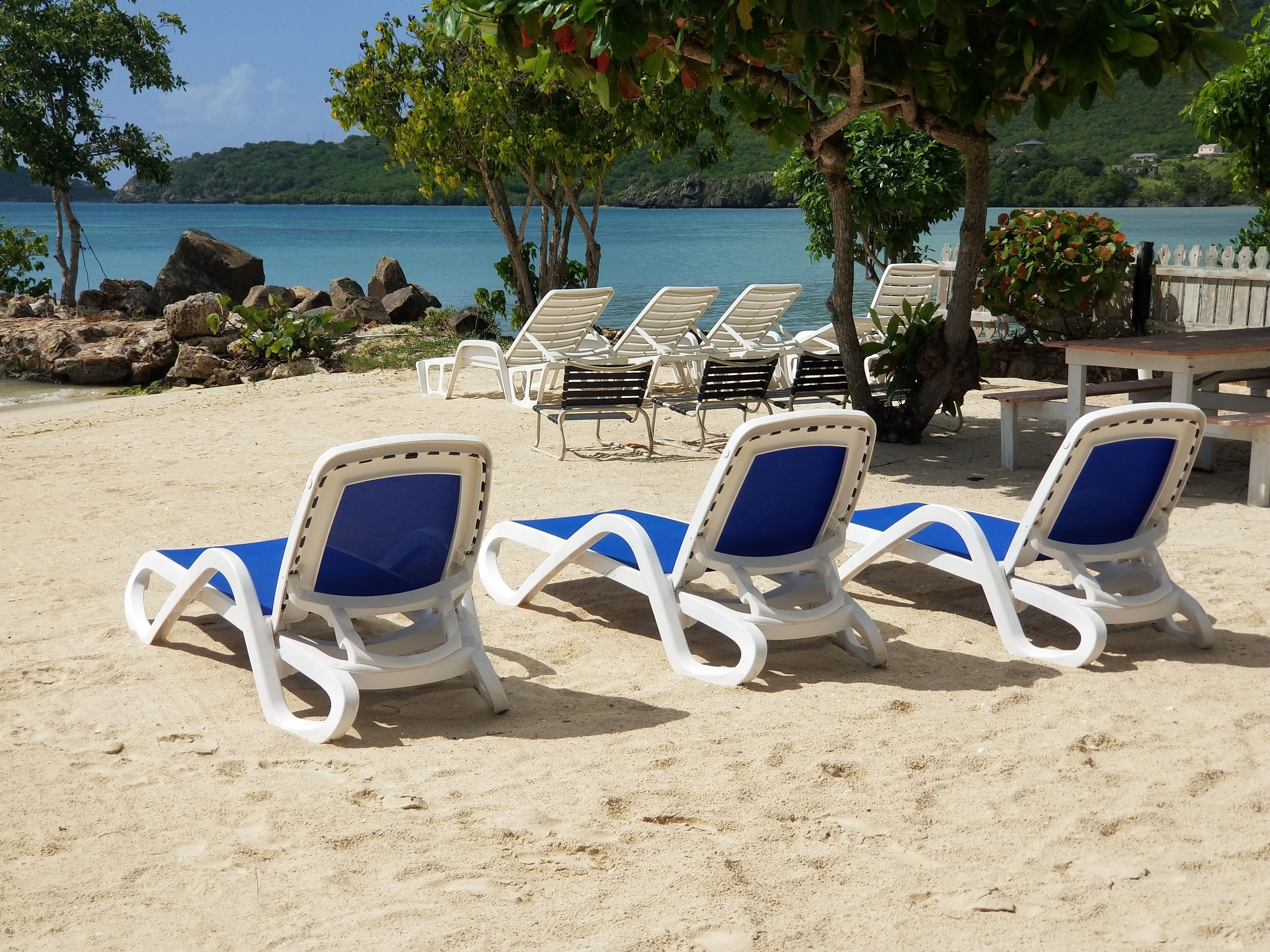 A beach with white lounge chairs with blue cushions, a wooden table, and a view of the sea and hills in the background.