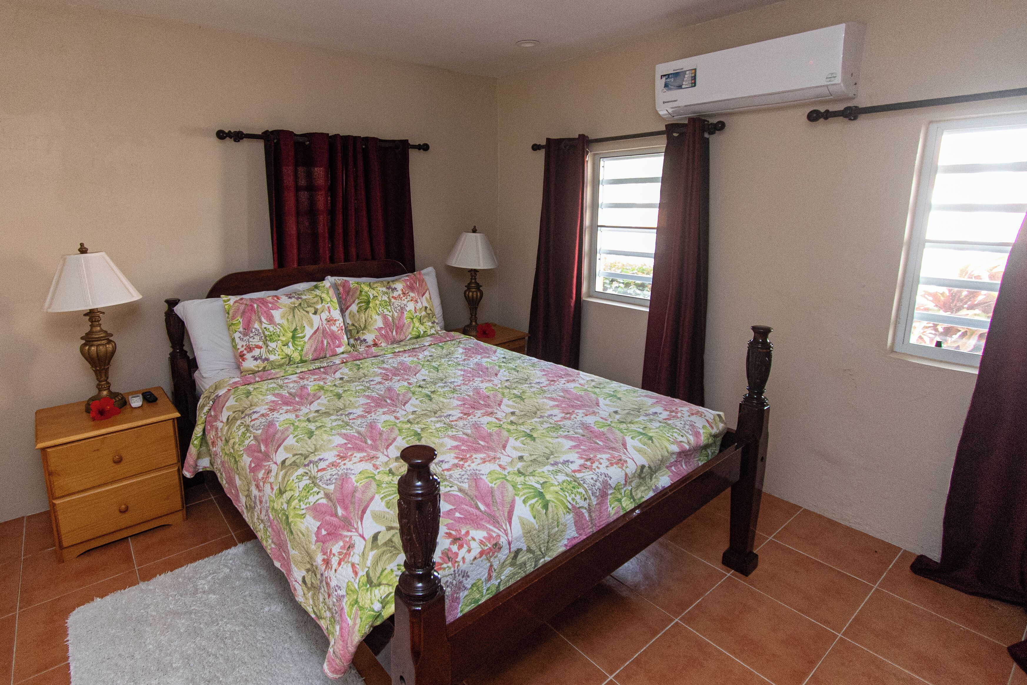 A bedroom with a bed, two bedside tables, a nightstand, a window with curtains, and an air conditioning unit.