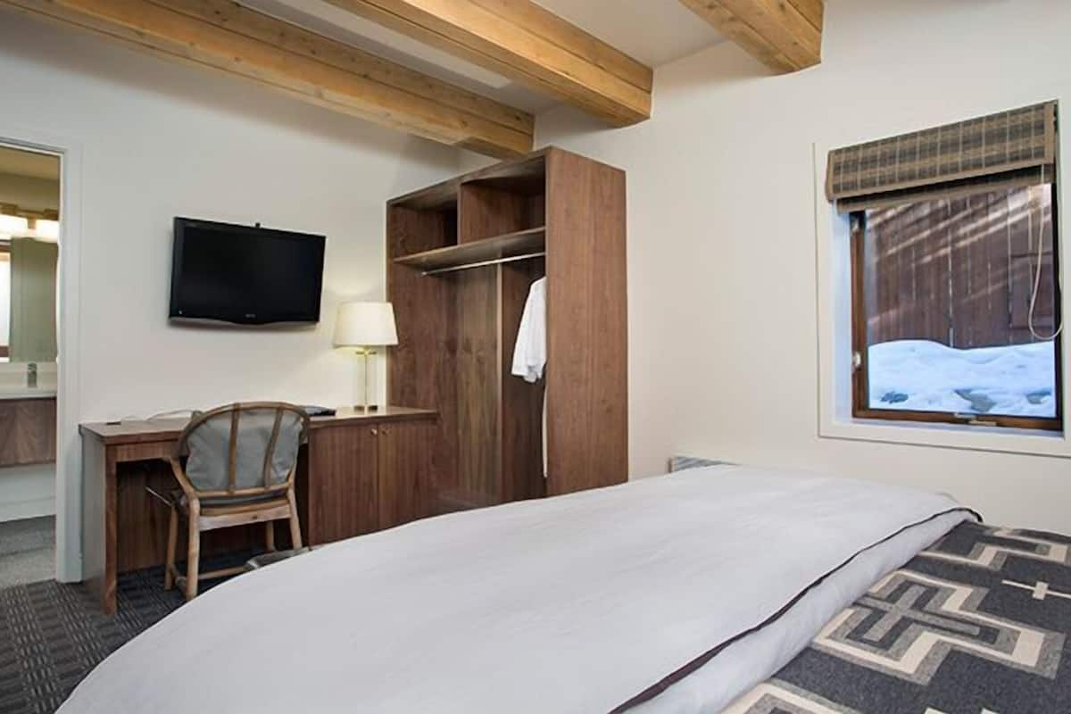 Habitación de hotel con cama, escritorio con silla, televisor, armario y ventana con vistas a la nieve.