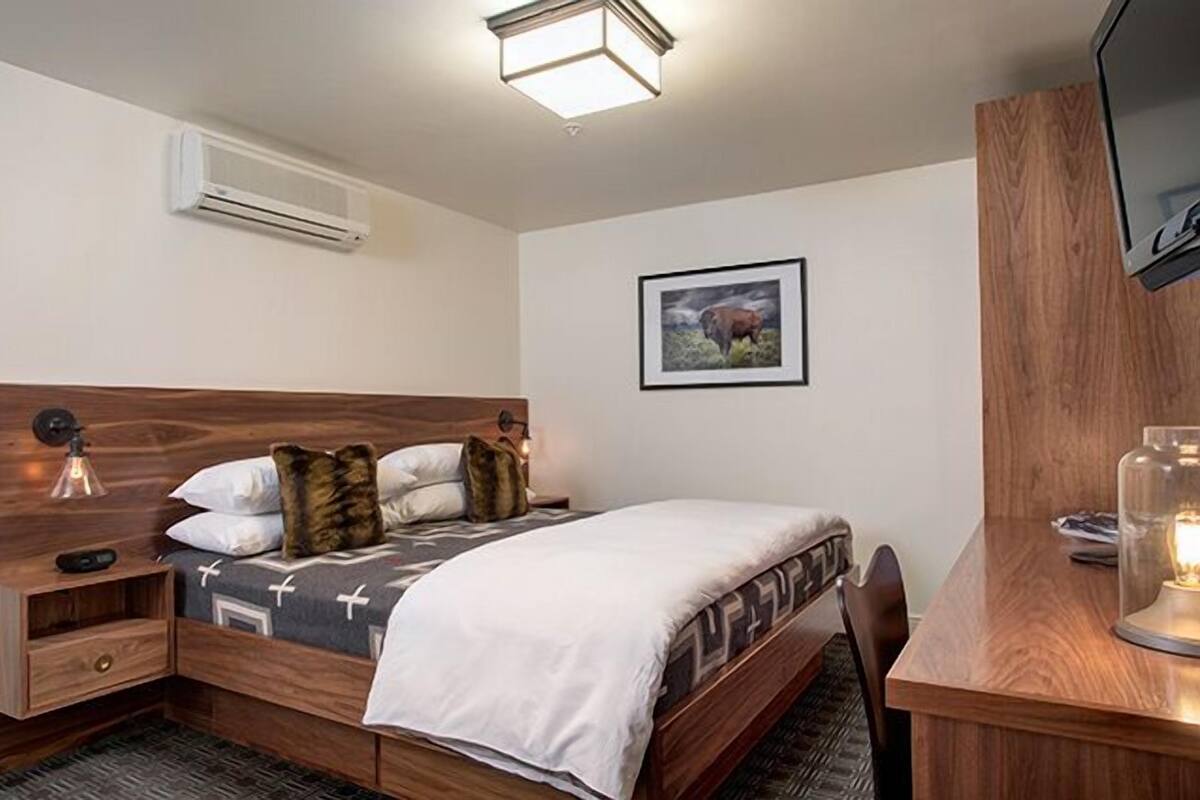 Un dormitorio con cabecera de madera, cama con ropa de cama estampada, mesita de noche con lámpara, escritorio con silla e imagen enmarcada en la pared.