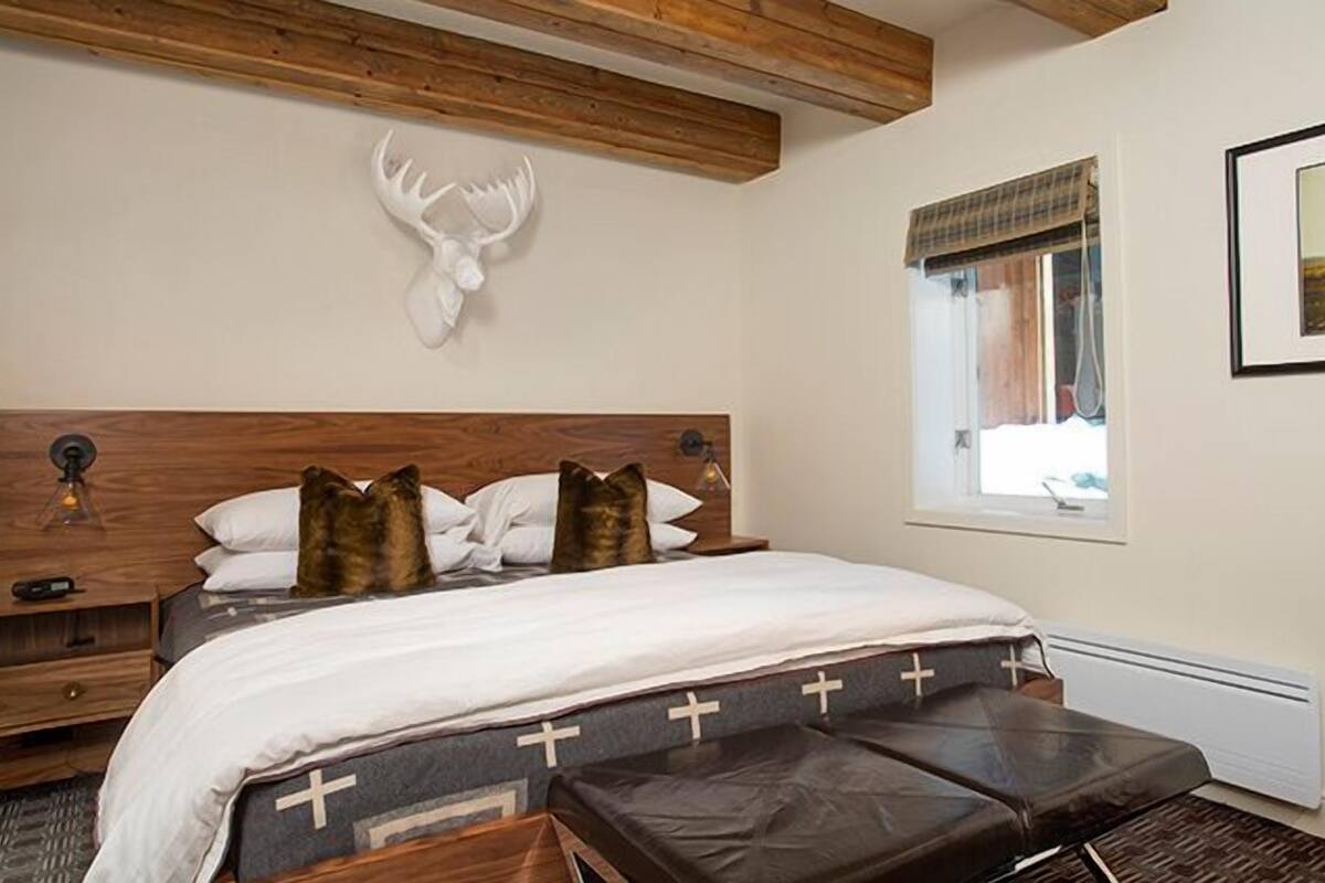 Un dormitorio con cabecera de madera, cráneo de alce montado en la pared, cama con ropa de cama blanca y colchón estampado en forma de cruz, y una ventana con vistas.