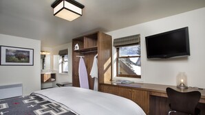 A hotel room with a bed, a wooden desk, a TV, and a window with a view of mountains. - Hotel Durant (Aspen)