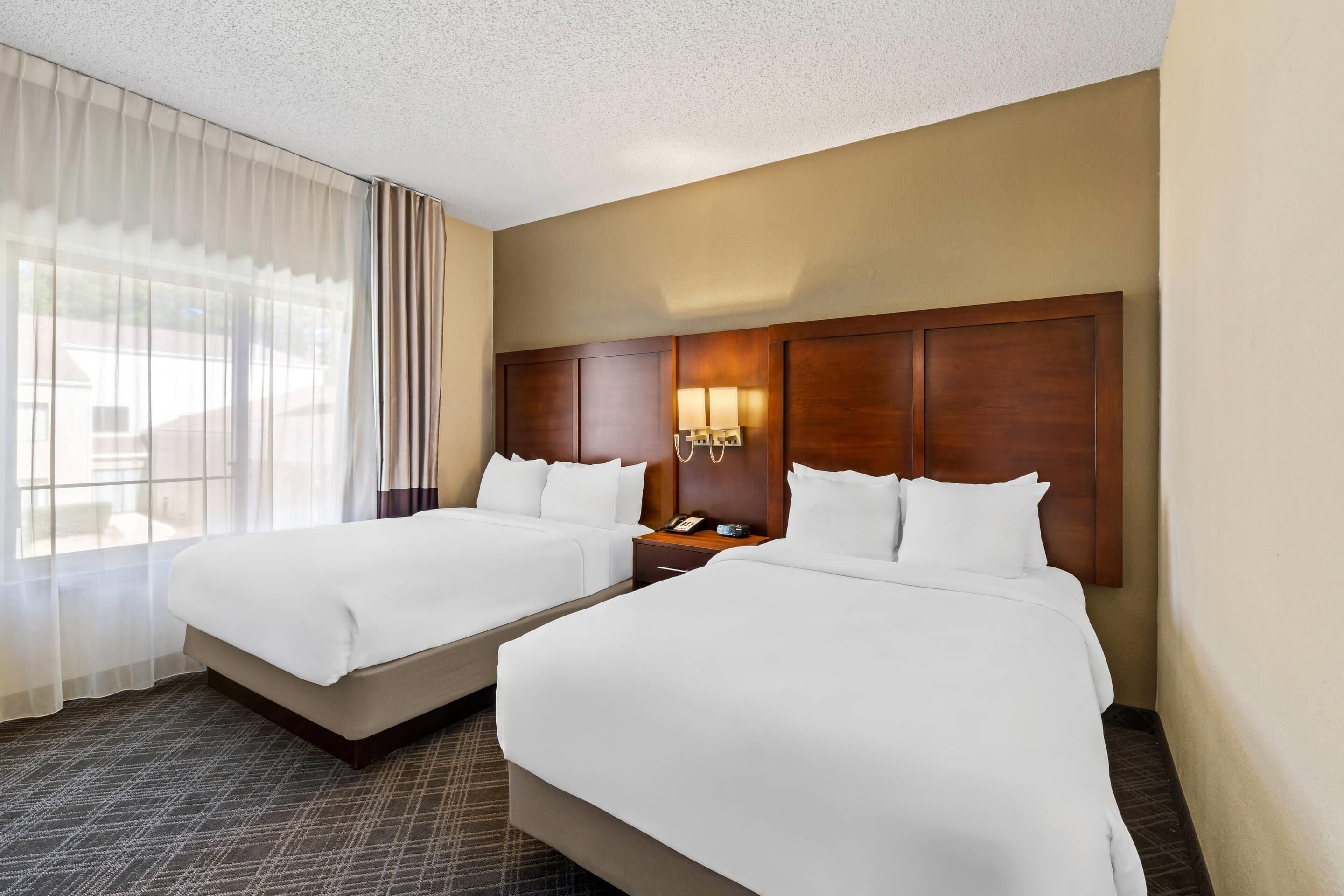 A hotel room with two beds, a wooden headboard, a nightstand with a lamp, and a window with sheer curtains.