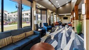 A modern hotel lobby with a large seating area, a patterned carpet, and large windows offering a view of the outdoor area.