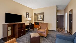 A hotel room with a flat-screen TV, a desk with a chair, a sofa, an ottoman, a mirror, and a door leading to another room.