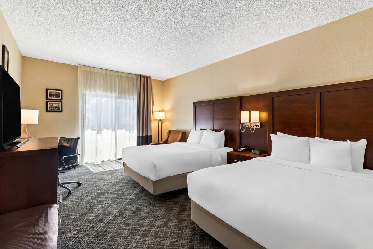 A hotel room with two beds, a desk, a chair, a lamp, and a TV.