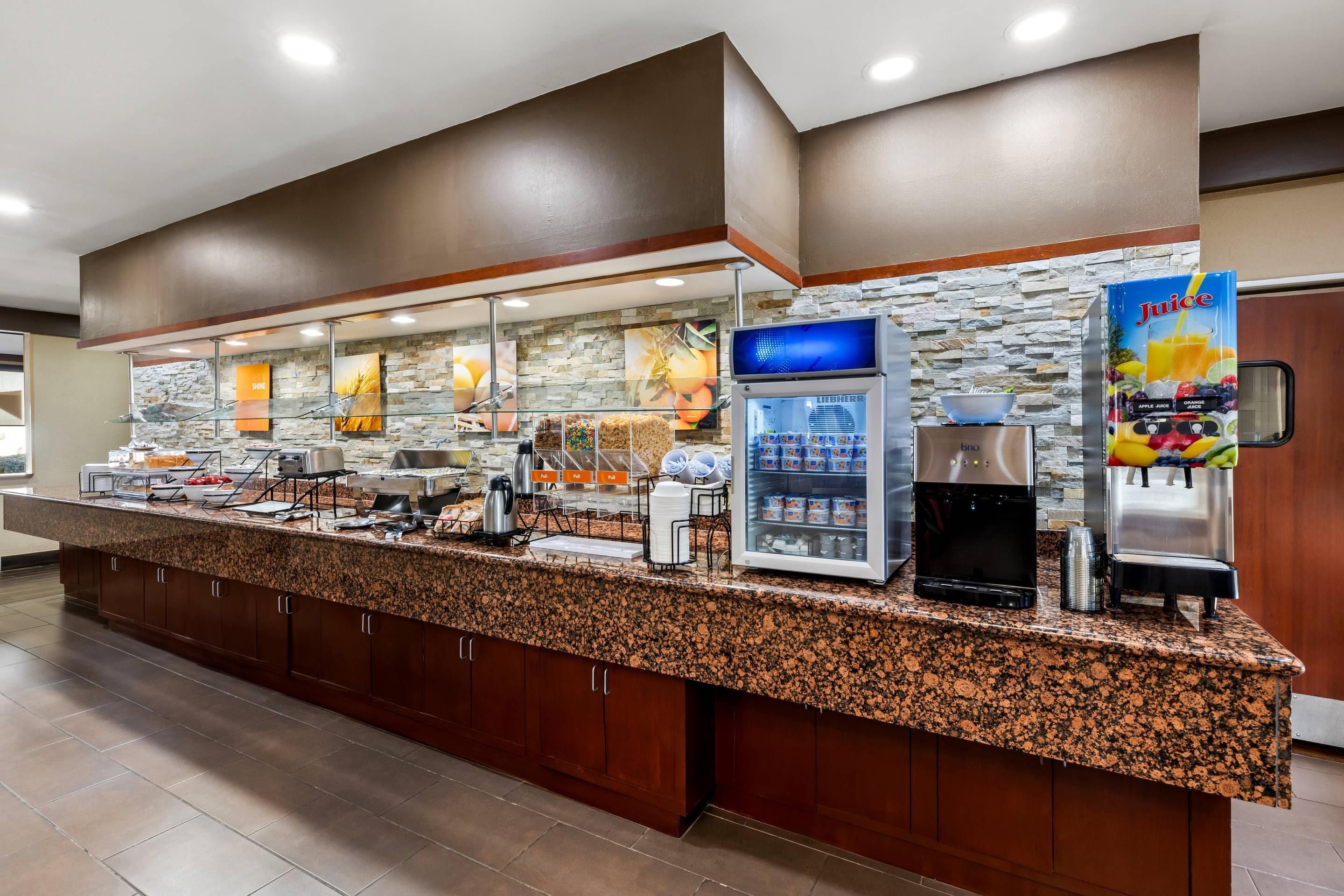 A self-service breakfast bar with a granite countertop, a coffee machine, a juice dispenser, and a refrigerated display case.