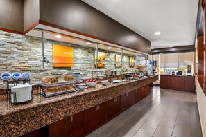 Uma área de buffet com parede de pedra ao fundo, um letreiro com a inscrição "RISE SHINE" e diversos alimentos expostos.
