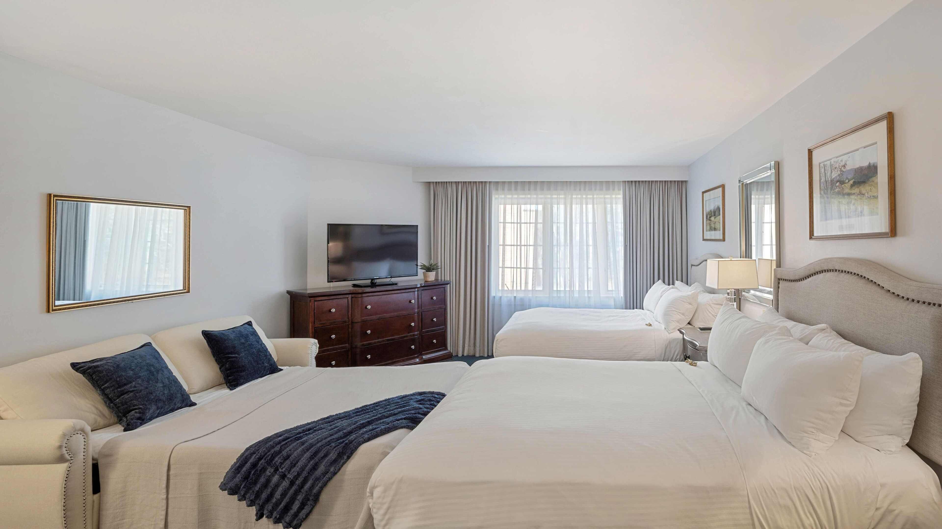 A hotel room with two beds, a TV, a dresser, and a window with curtains.