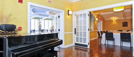 A room with a piano, a dining area with a table and chairs, and an exit sign on the wall.
