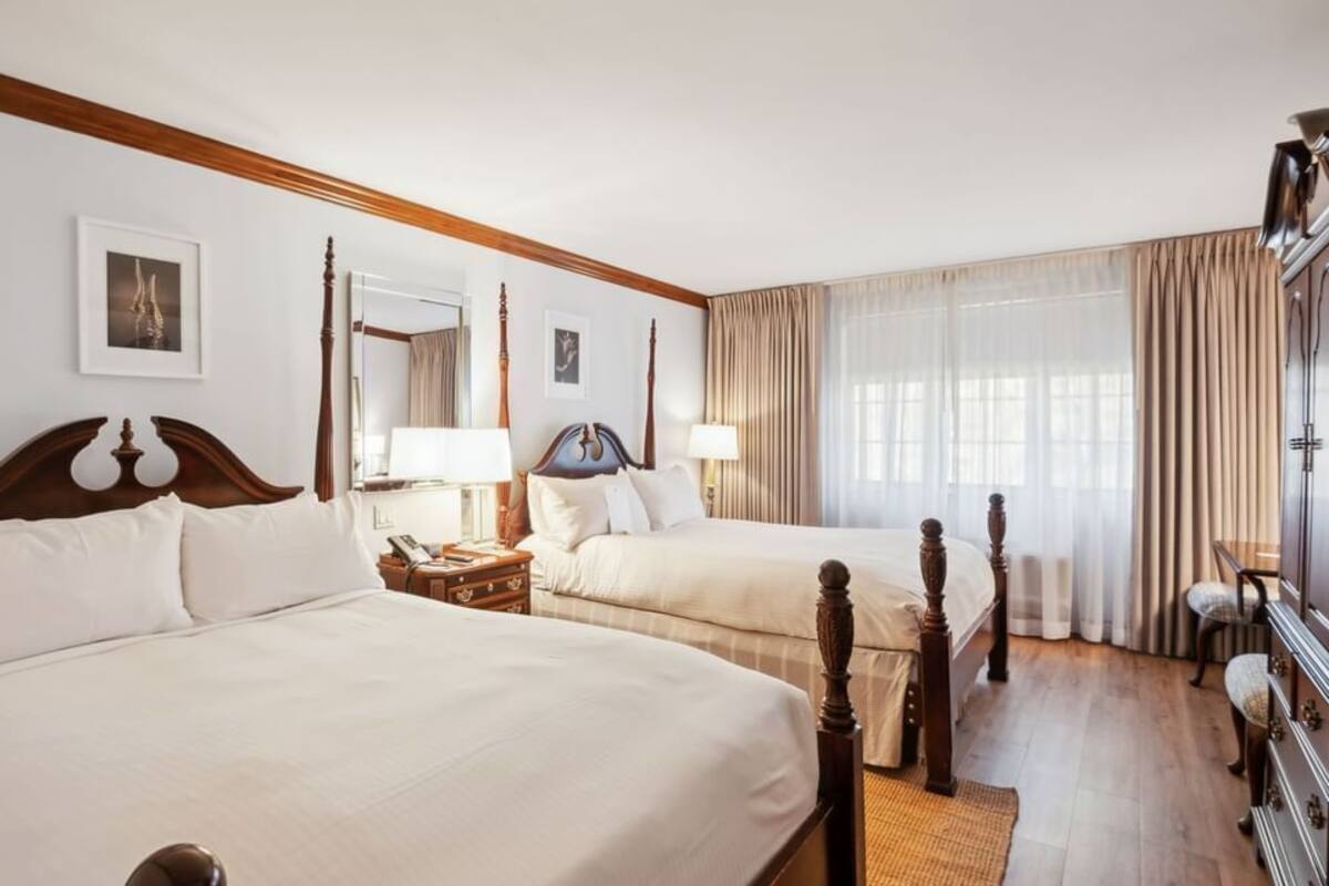 A hotel room with two large beds, wooden headboards, a nightstand, a dresser, and a large window with curtains.