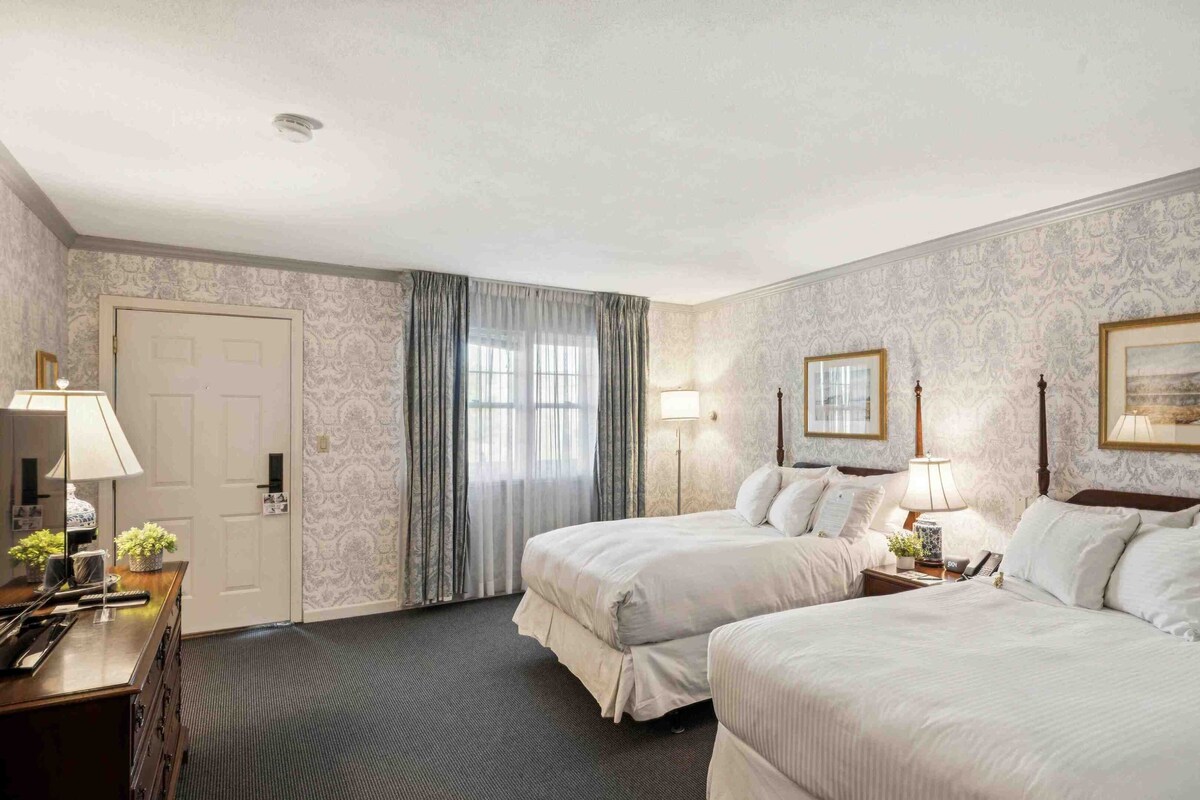 A hotel room with two beds, a desk with a lamp, a chair, and a window with curtains.