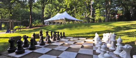 A large outdoor chessboard set up on a grassy area with a pavilion in the background.
