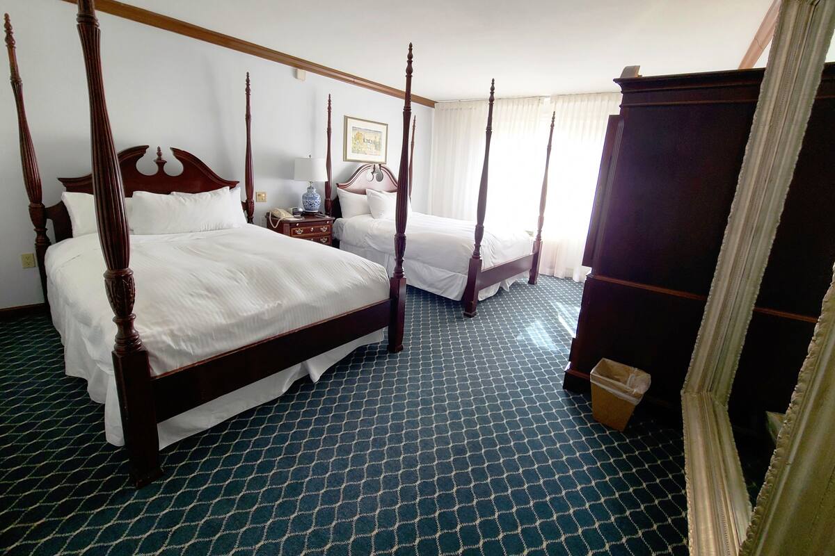 A four-poster bed with white linens, a bedside table with a lamp, and a patterned carpet.