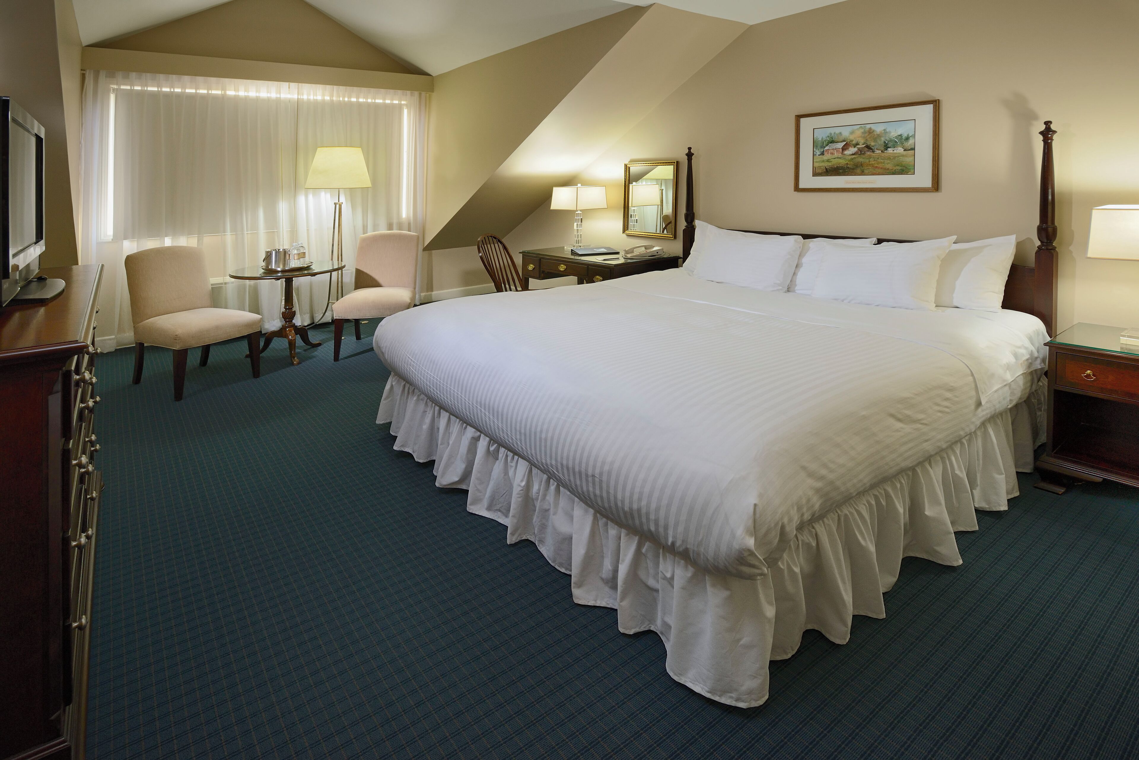 Une chambre d’hôtel avec un grand lit, deux chaises, un bureau, une lampe et un tableau au mur.