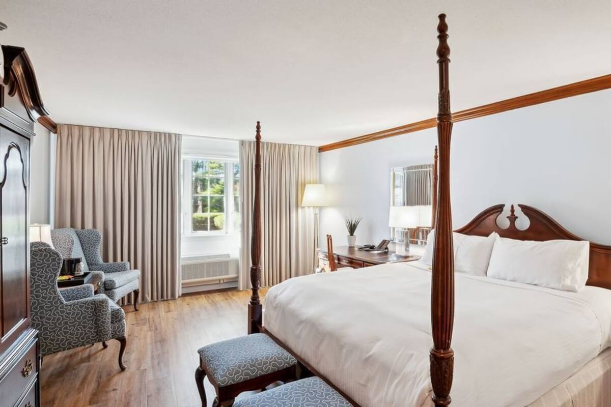 A four-poster bed with a wooden headboard, a sitting area with a chair and ottoman, a desk with a lamp, and a large mirror on the wall.