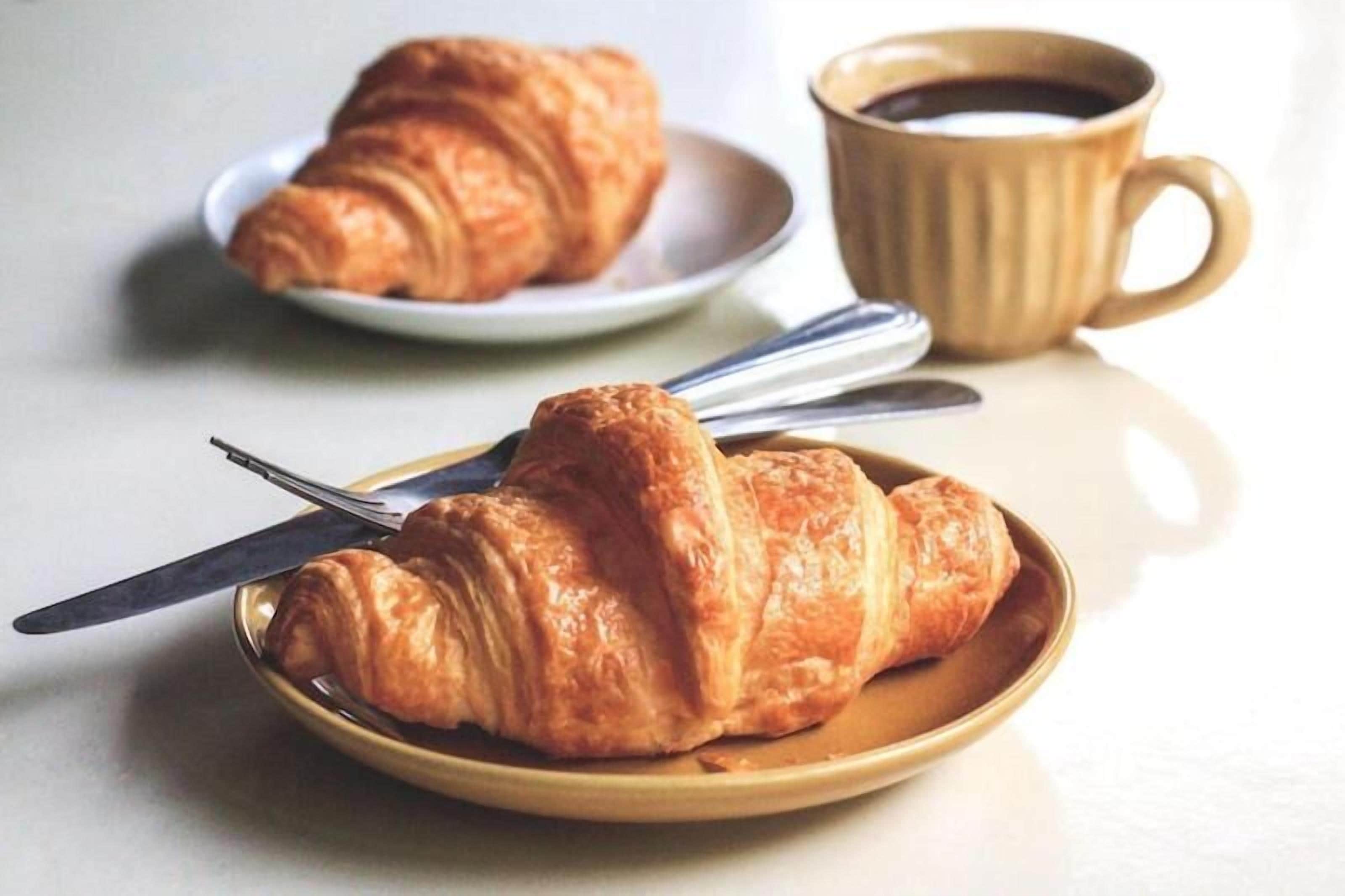 Zwei Croissants auf Tellern, eines mit einem Messer, und eine Tasse Kaffee auf einem Tisch.