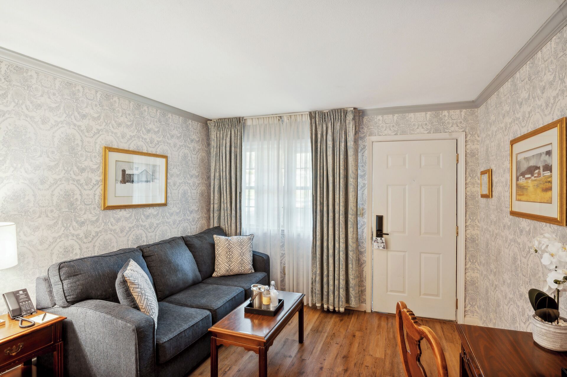 A living room with a dark grey sofa, a wooden coffee table, a desk with a telephone, and a door with a white frame.