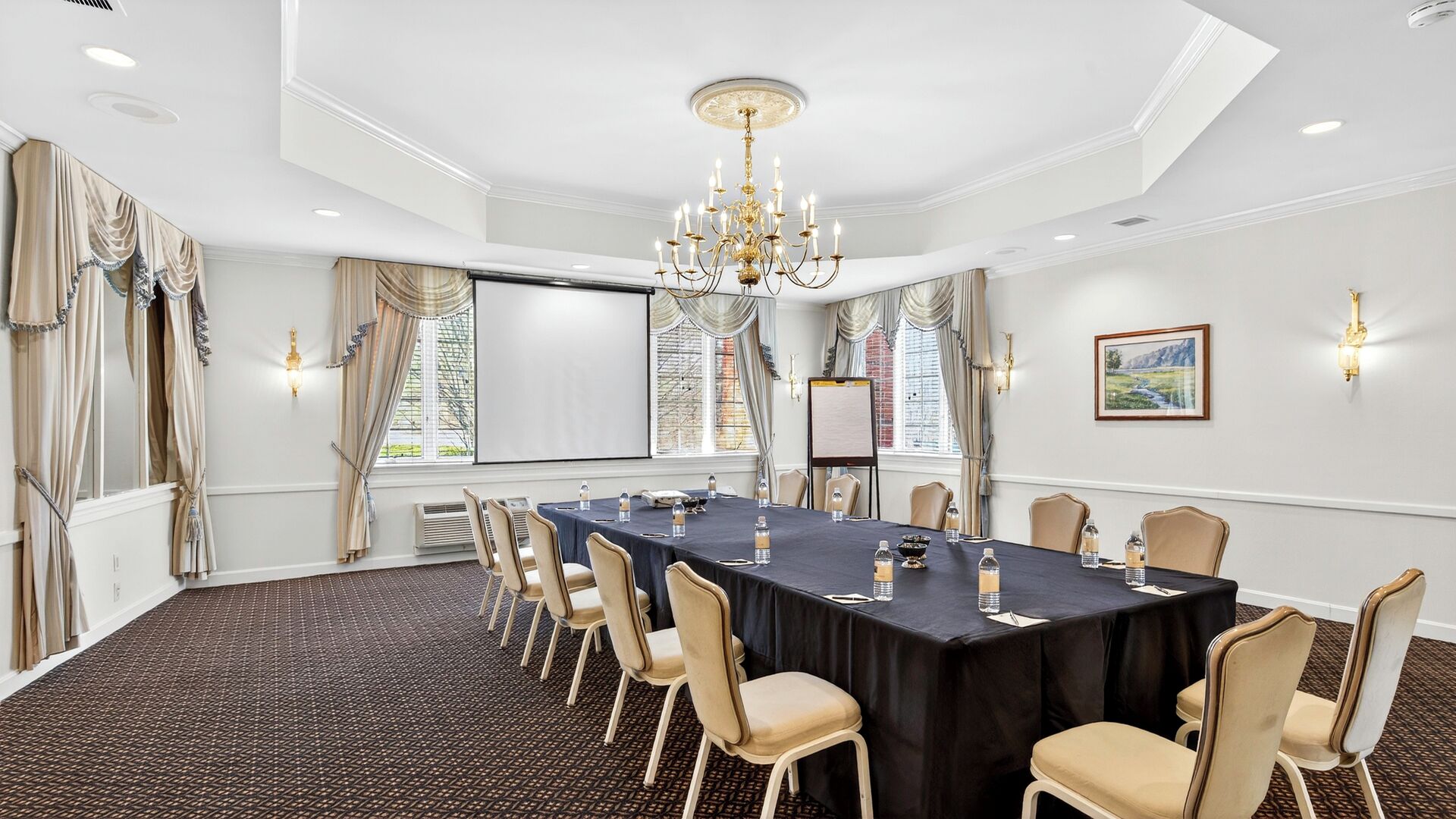 A conference room with a long table, chairs, a chandelier, windows with curtains, and a projector screen.