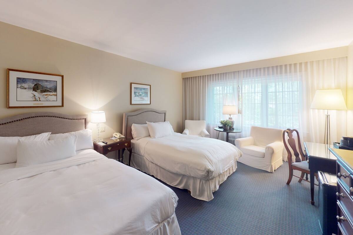 A hotel room with two beds, a desk, a chair, and a lamp.