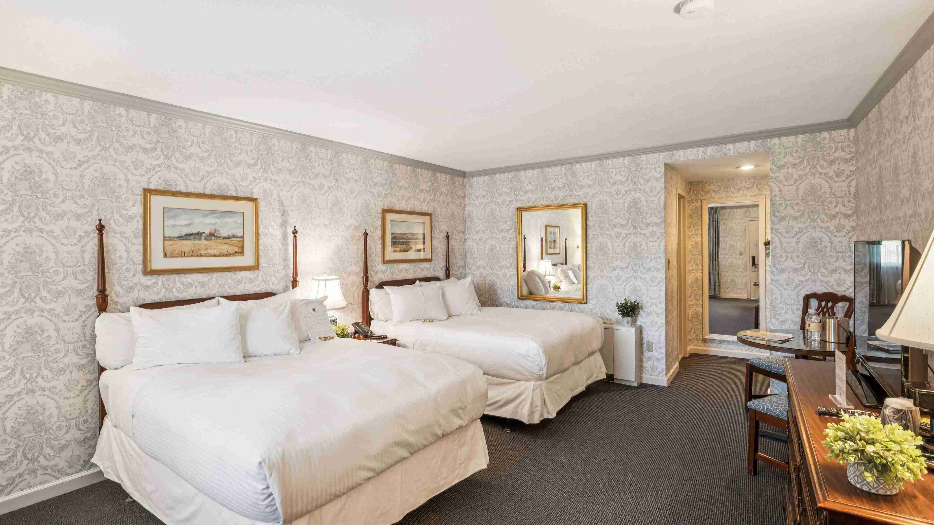 A hotel room with two beds, a desk, a chair, a mirror, and a small table with a lamp.
