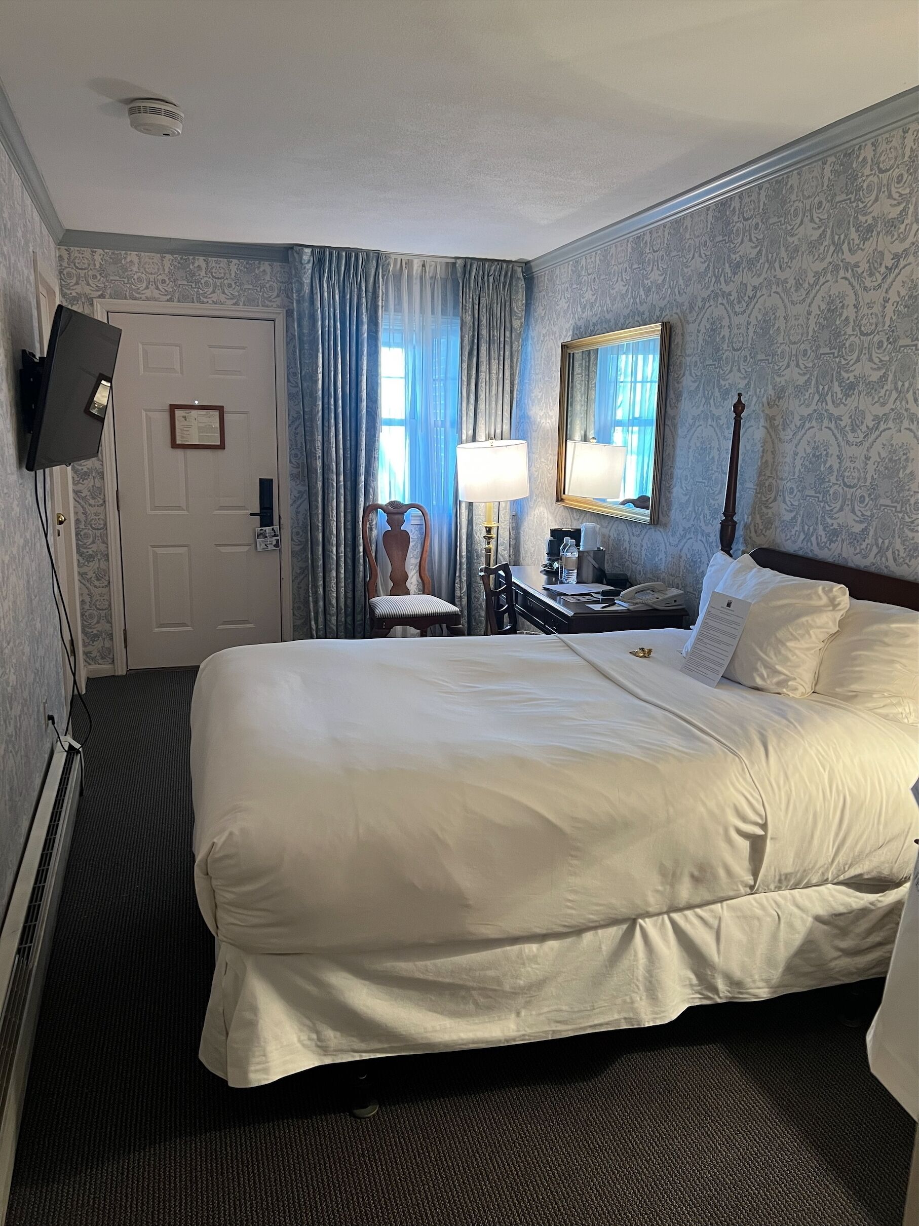 A hotel room with a large bed, a desk with a lamp, a chair, a television mounted on the wall, and a window with curtains.