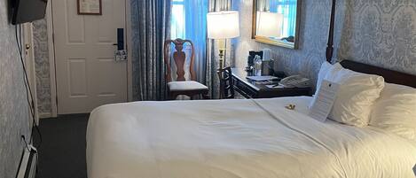 Habitación de hotel con una cama grande, un escritorio con lámpara, una silla, un televisor montado en la pared y una ventana con cortinas.