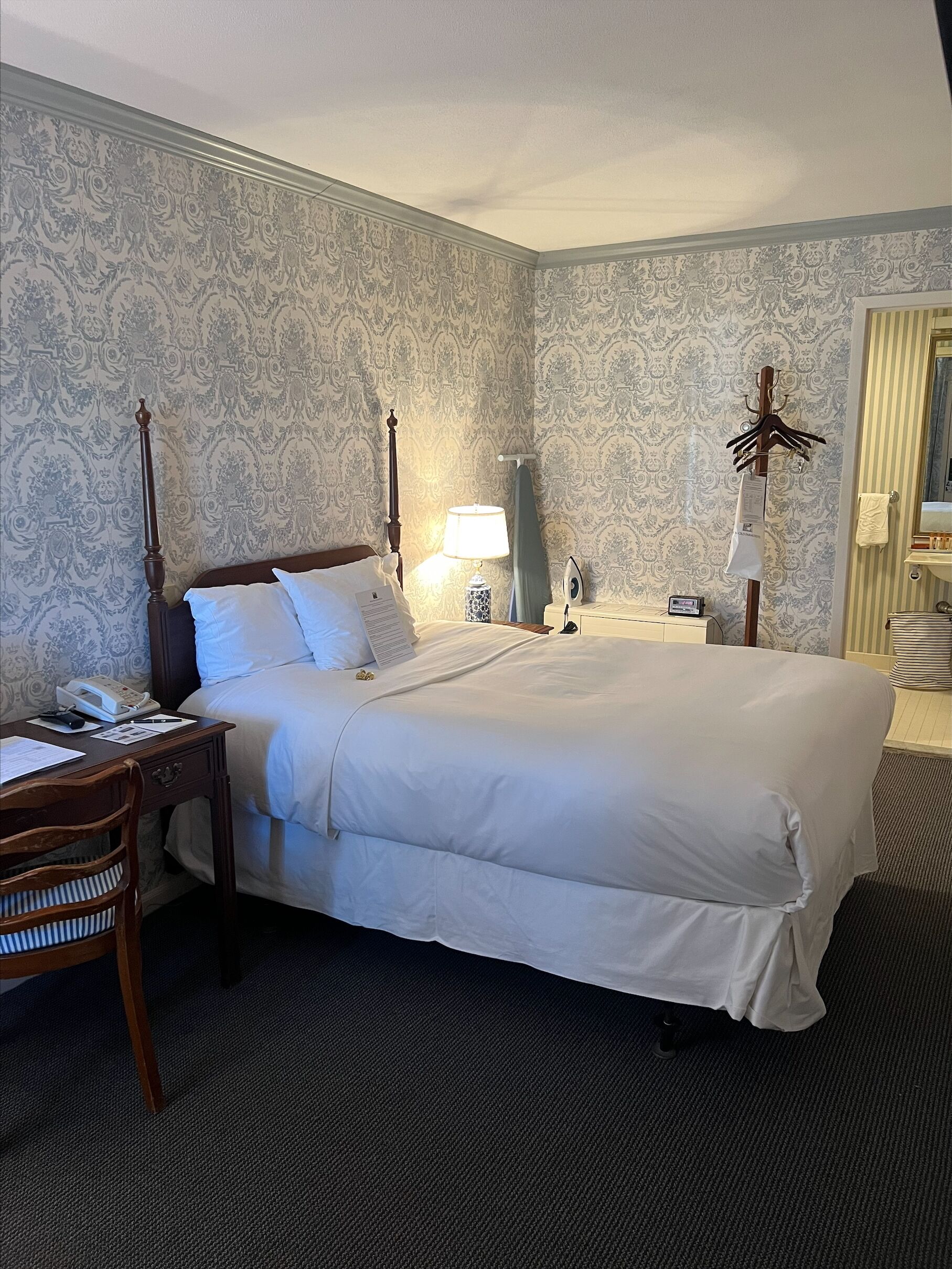 A hotel room with a large bed, a desk with a telephone, a chair, a bedside table with a lamp, and a mirror.